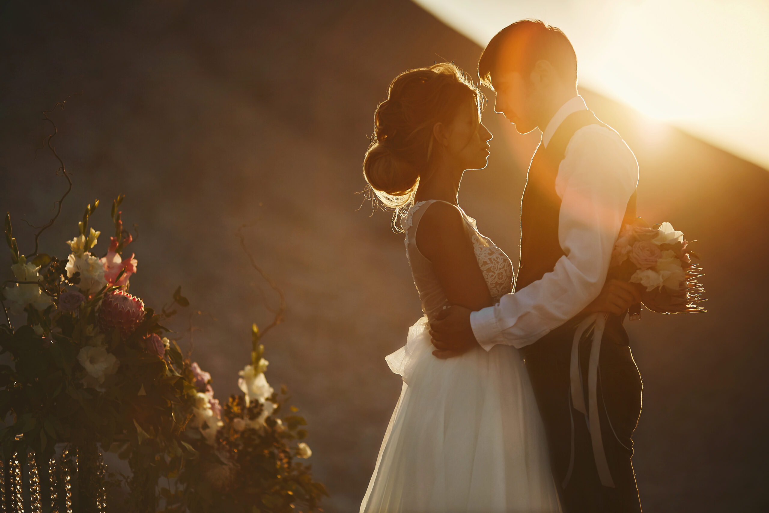 Silhouette sweethearts at sunset. Beautiful and fashionable brunette model girl with modeling hairstyle in white lace dress posing at the sunset with handsome men in stylish vest and white shirt.
